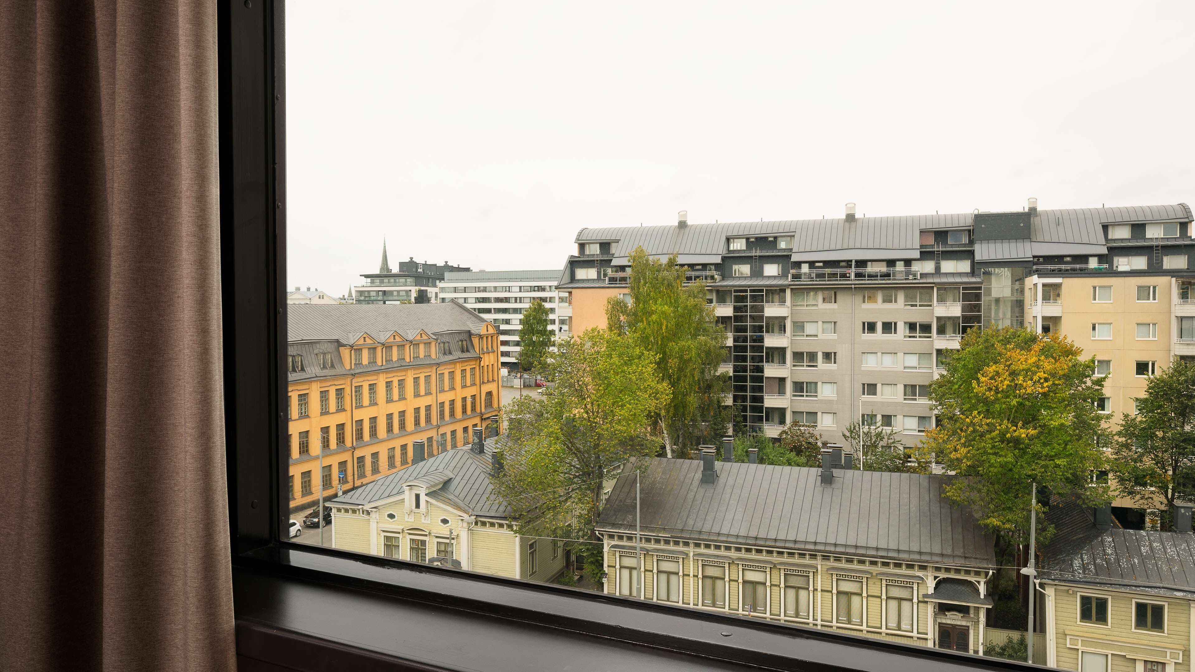 Une vue sur un paysage urbain à travers une fenêtre avec des rideaux, mettant en scène des immeubles de plusieurs étages aux styles architecturaux variés, notamment un bâtiment jaune au design classique et des gratte-ciel modernes.