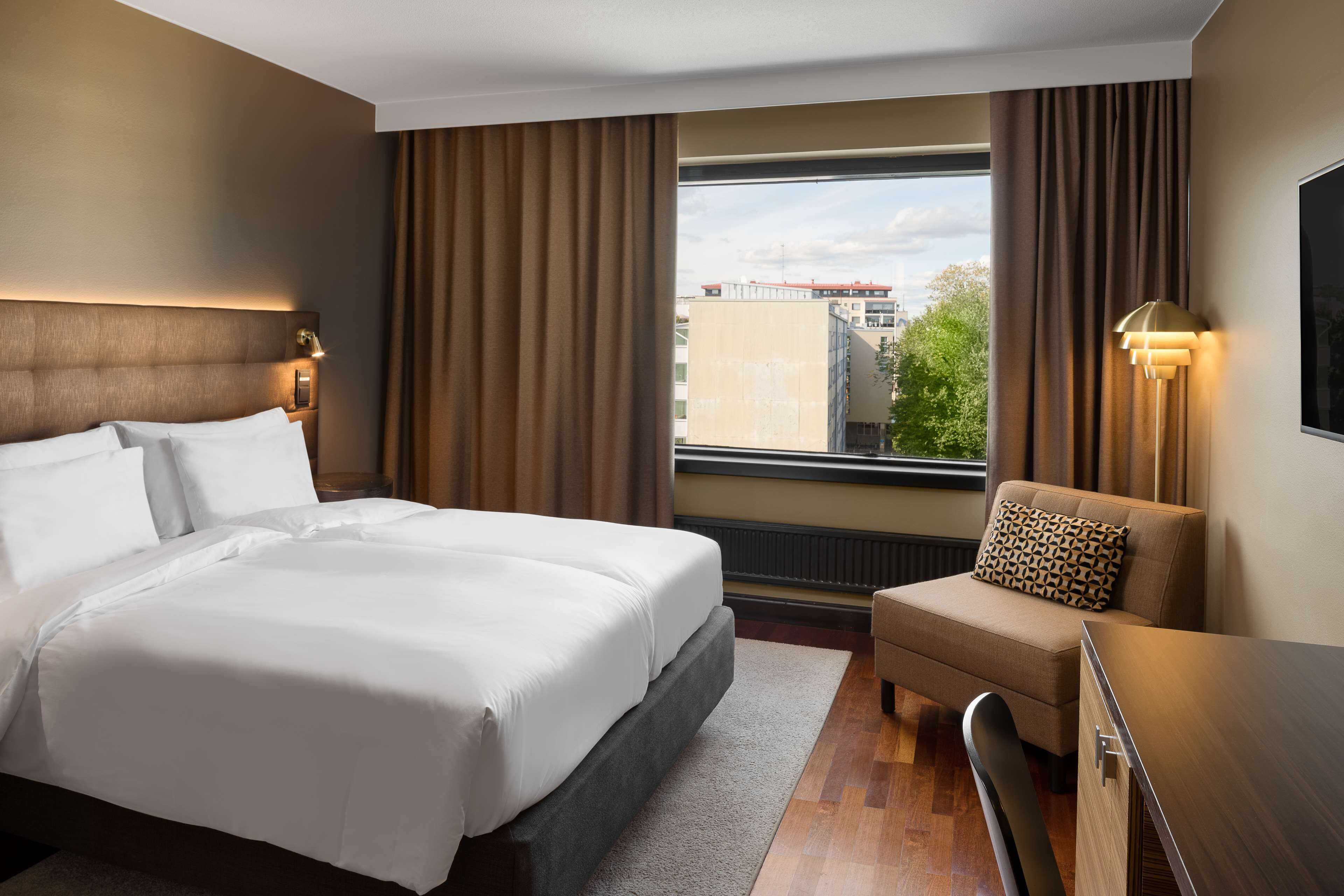 Une chambre d’hôtel avec un grand lit, un canapé, un bureau et une fenêtre donnant sur des immeubles et des arbres.