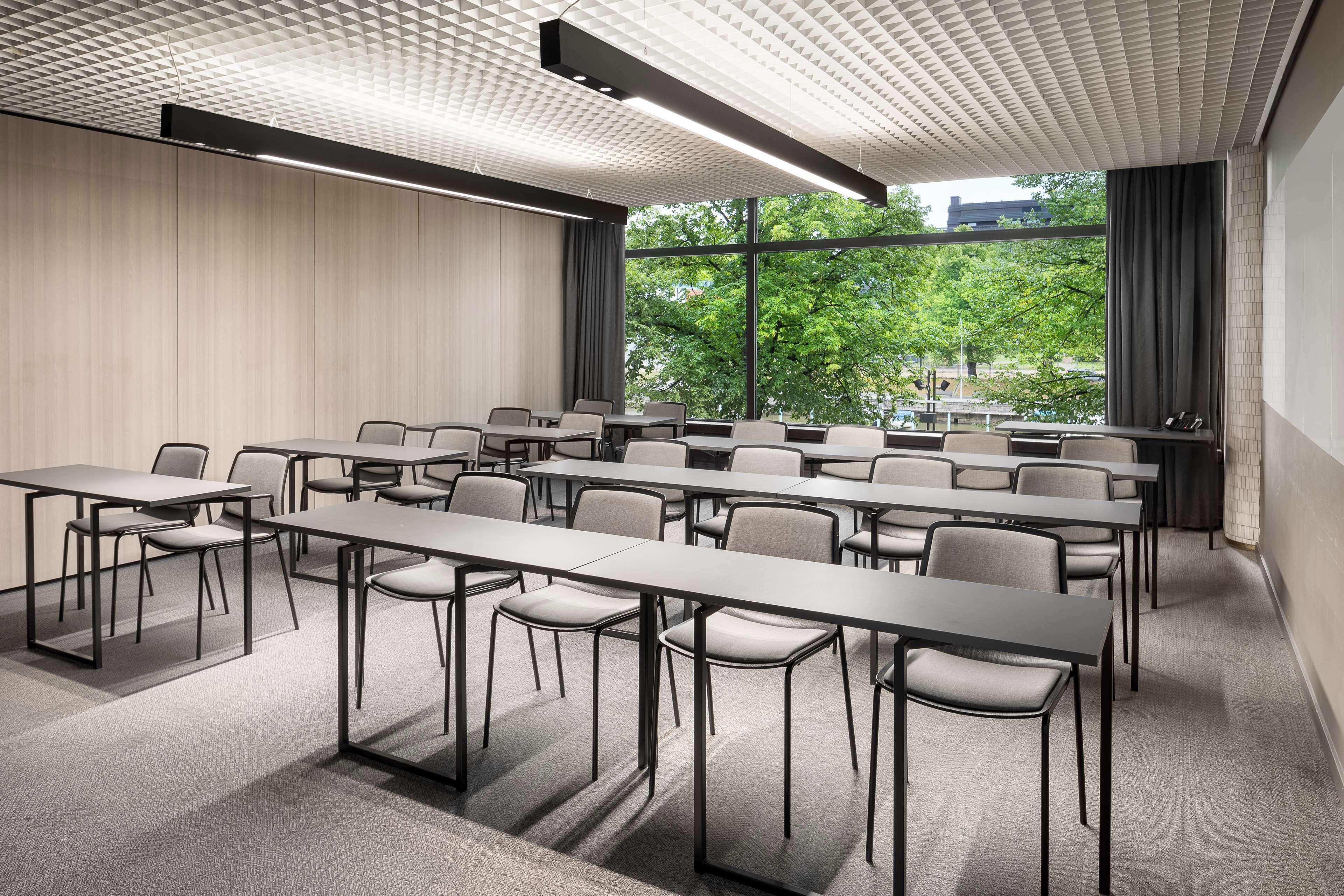 Une salle de conférence dotée de plusieurs tables et chaises, de grandes fenêtres avec des rideaux et d’un plafond orné d’un motif géométrique.