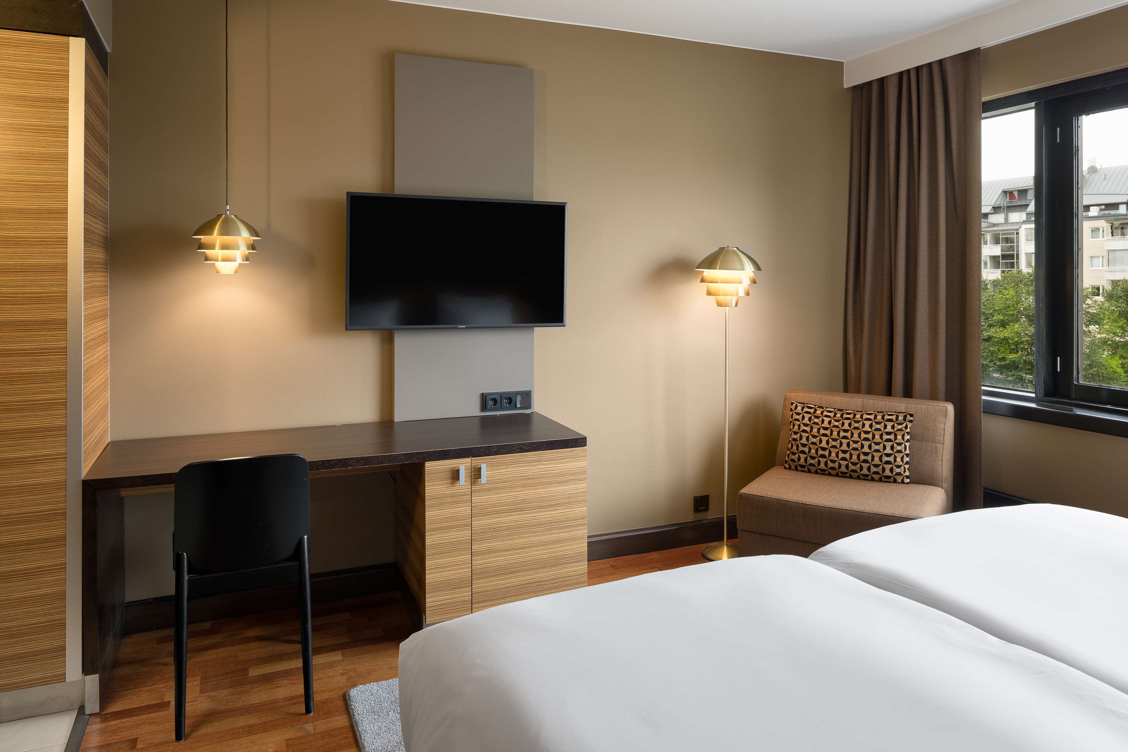 A hotel room with a bed, a desk with a chair, a TV mounted on the wall, and a window with curtains.