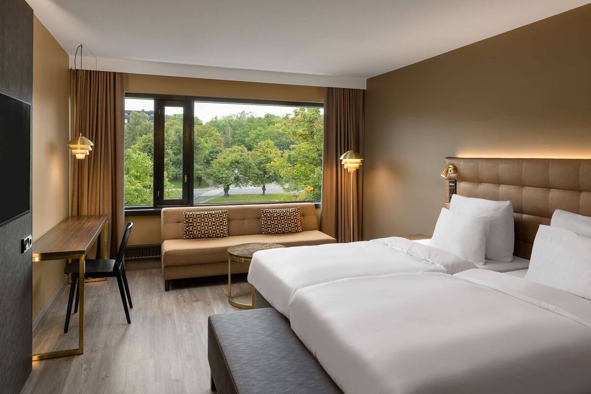 Ein Hotelzimmer mit einem großen Bett, einem Sofa, einem Schreibtisch und Blick auf Bäume aus dem Fenster.
