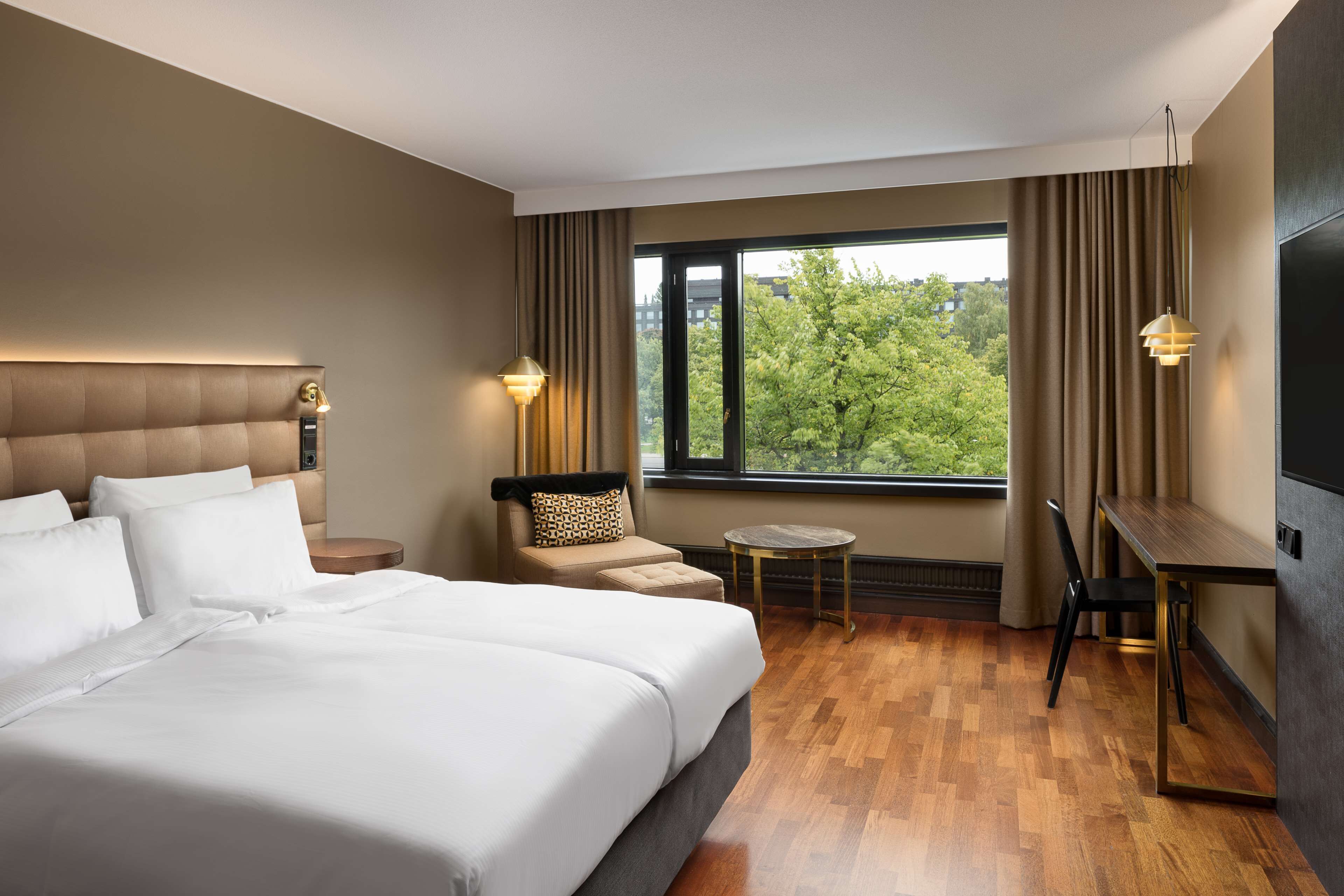 Hotellihuone, jossa on suuri sänky, työpöytä, tuoli, pieni pyöreä pöytä ja ikkunasta avautuva näkymä puita kohti.