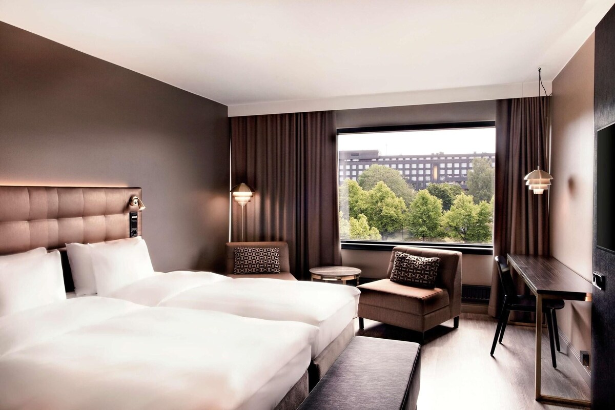 A hotel room with a large bed, a desk, a chair, and a view of trees and buildings through the window.