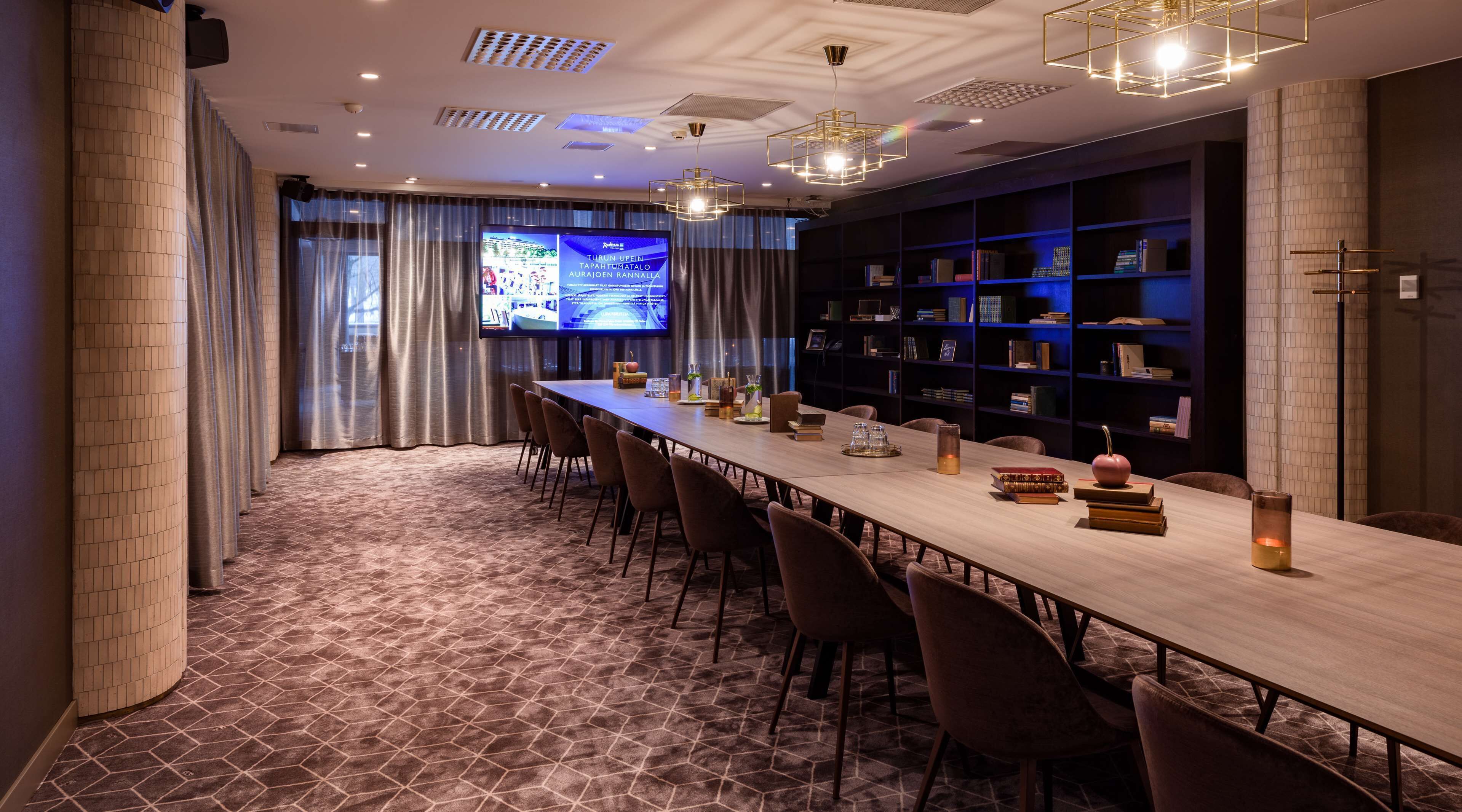 Un salón de conferencias con una larga mesa, sillas, una pantalla de televisión y estanterías.