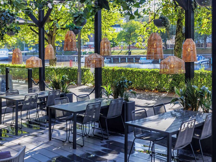 An outdoor dining area with tables and chairs, surrounded by greenery and hanging wicker lamps.
