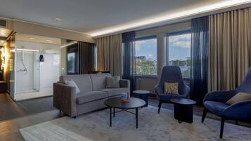 Habitación de hotel moderna con sofá gris, dos sillones, una mesita de centro y ducha con cerramiento de vidrio. Amplias ventanas ofrecen vistas a la ciudad.