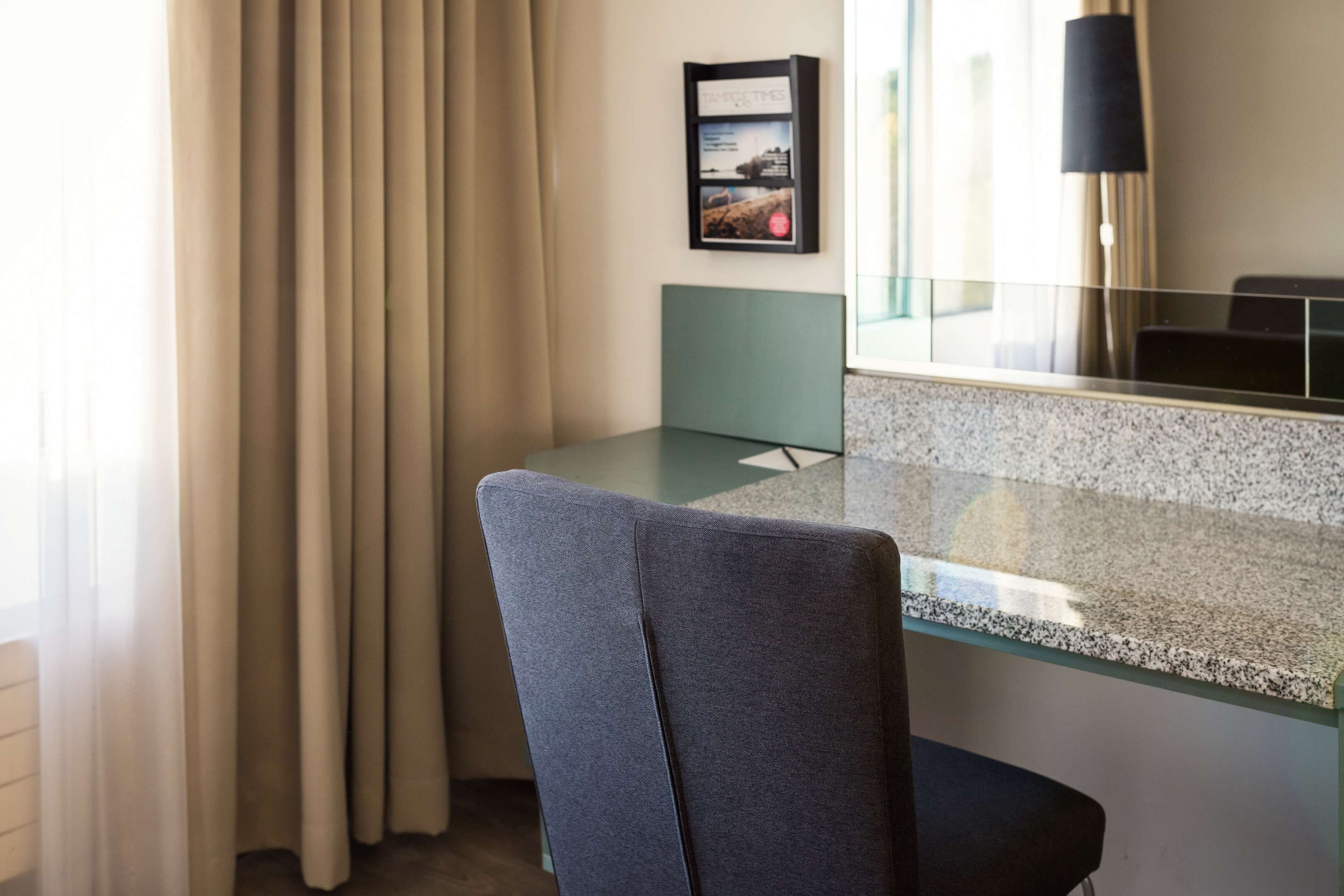 Ein Hotelzimmer mit Glastisch, grauem Stoffstuhl, einem gerahmten Bild an der Wand und einem Fenster mit Vorhängen.