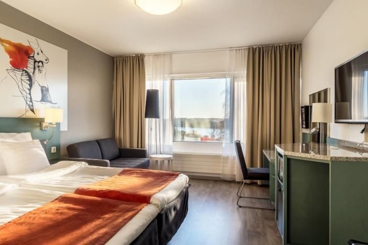 Habitación de hotel con cama, escritorio, silla, televisor y ventana con cortinas.