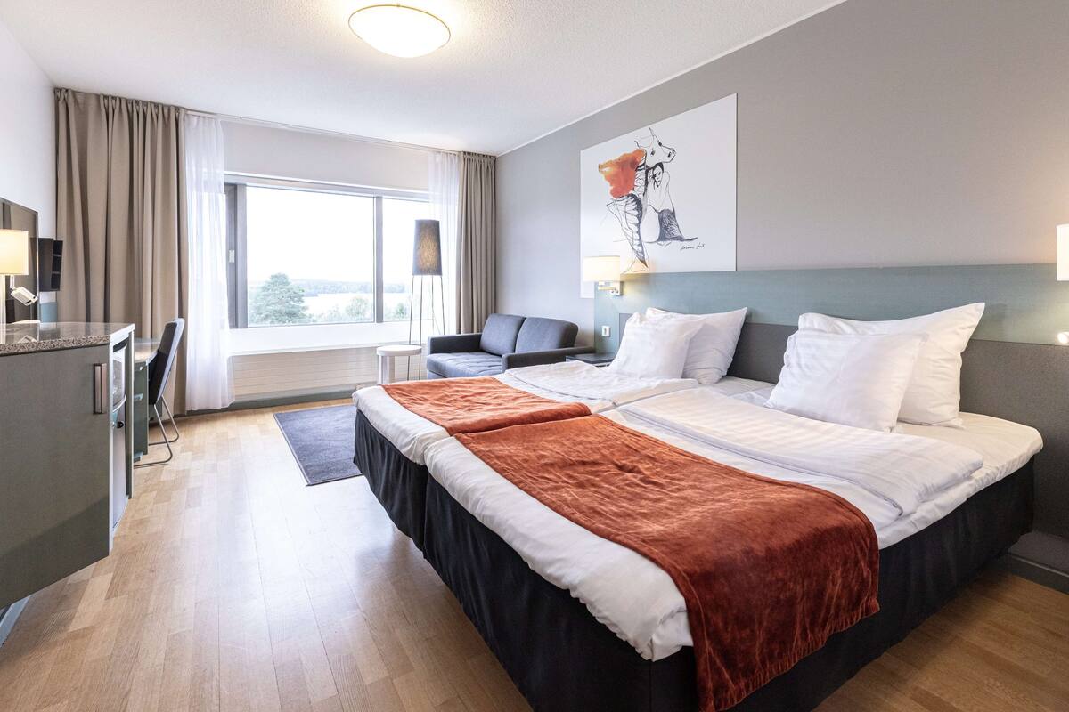 A hotel room with a bed, a desk, a chair, a window with curtains, and a painting on the wall.