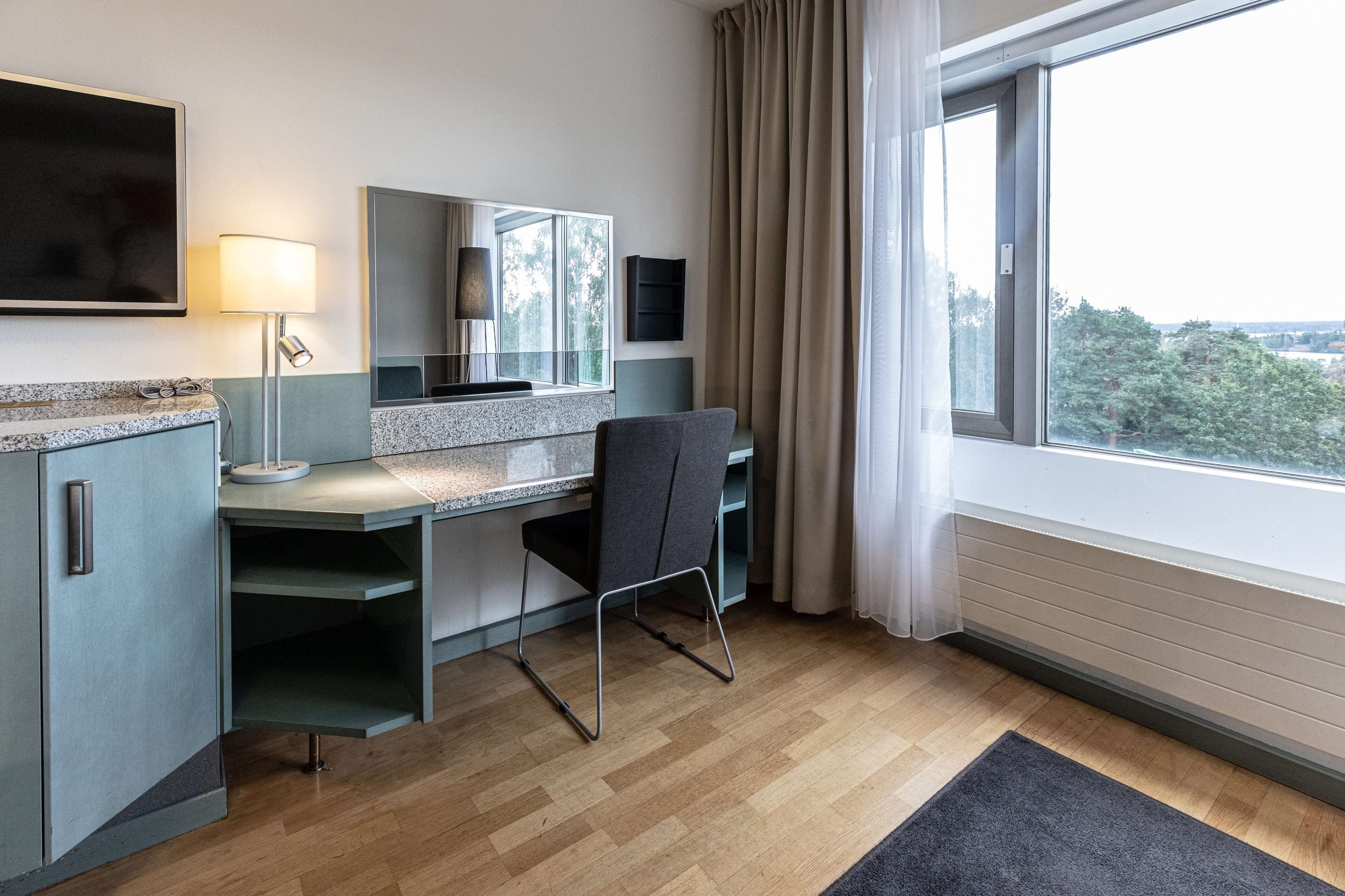 A modern hotel room with a desk, chair, and large window offering a view of the outdoors.