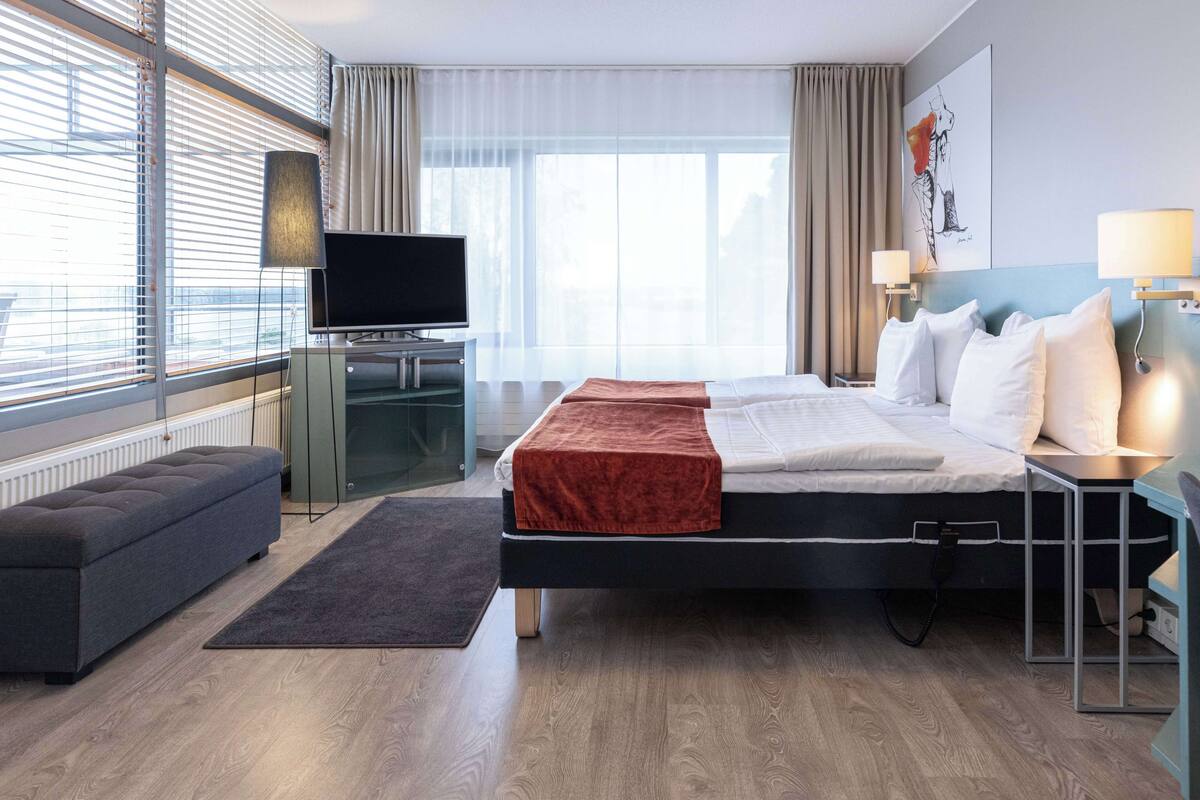A modern hotel room with a large bed, a desk with a TV, a sofa, and a view of the city through large windows.