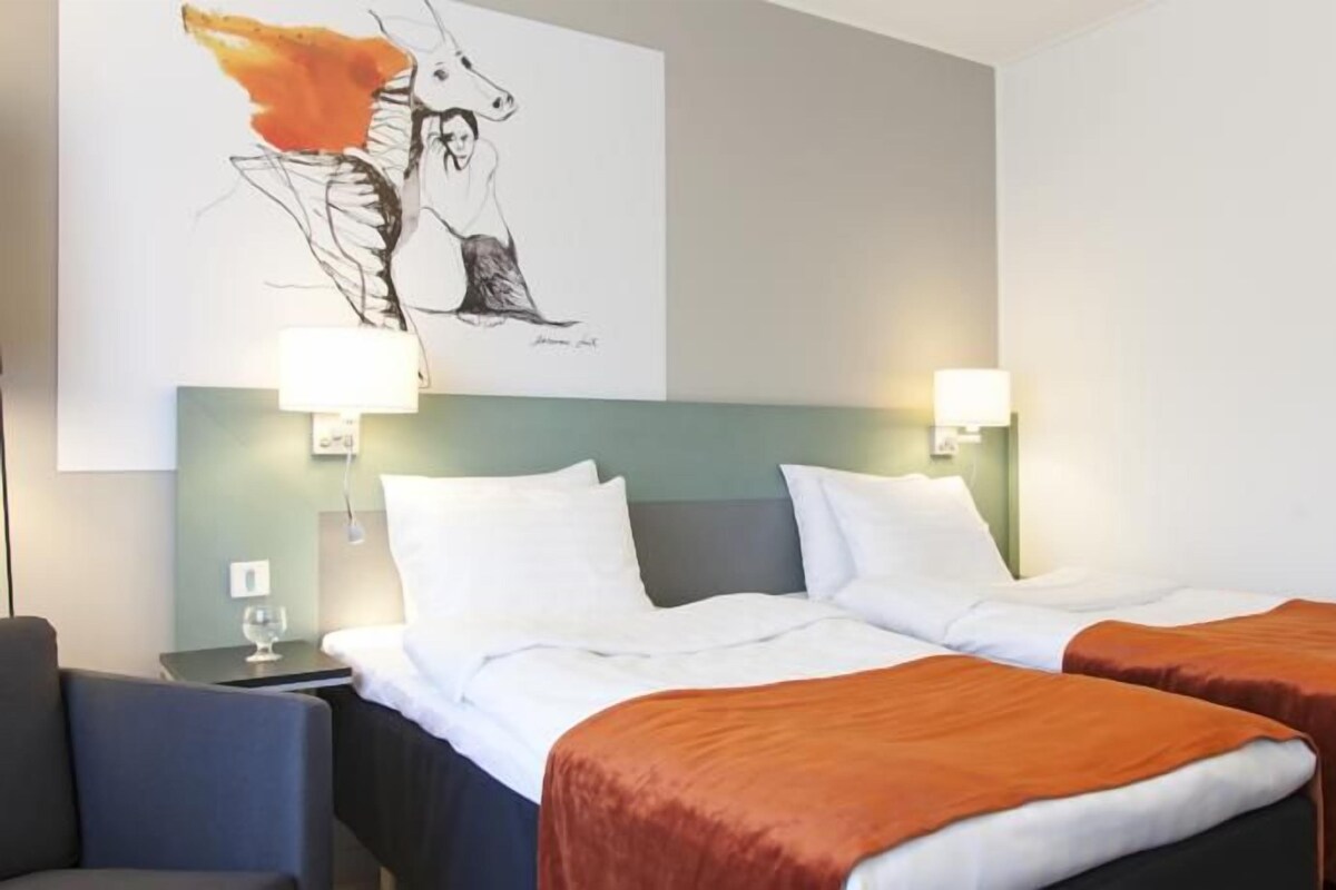 Habitación de hotel con dos camas, una mesita de noche, una lámpara y una obra de arte colgada en la pared.