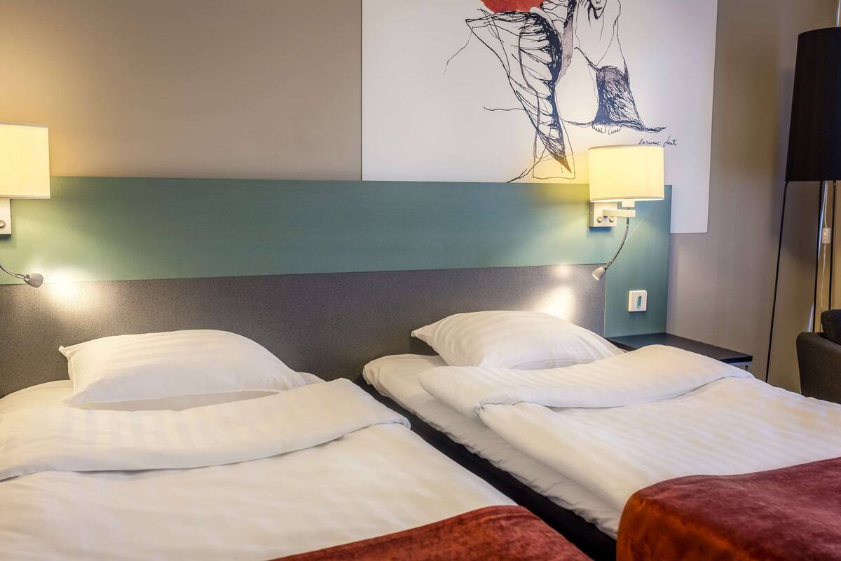 A hotel room with two beds, a headboard, wall-mounted lamps, and a painting.
