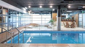 Une piscine intérieure avec des parois en verre transparent, un espace détente avec des fauteuils et un mur en pierre agrémenté de plantes en pot.