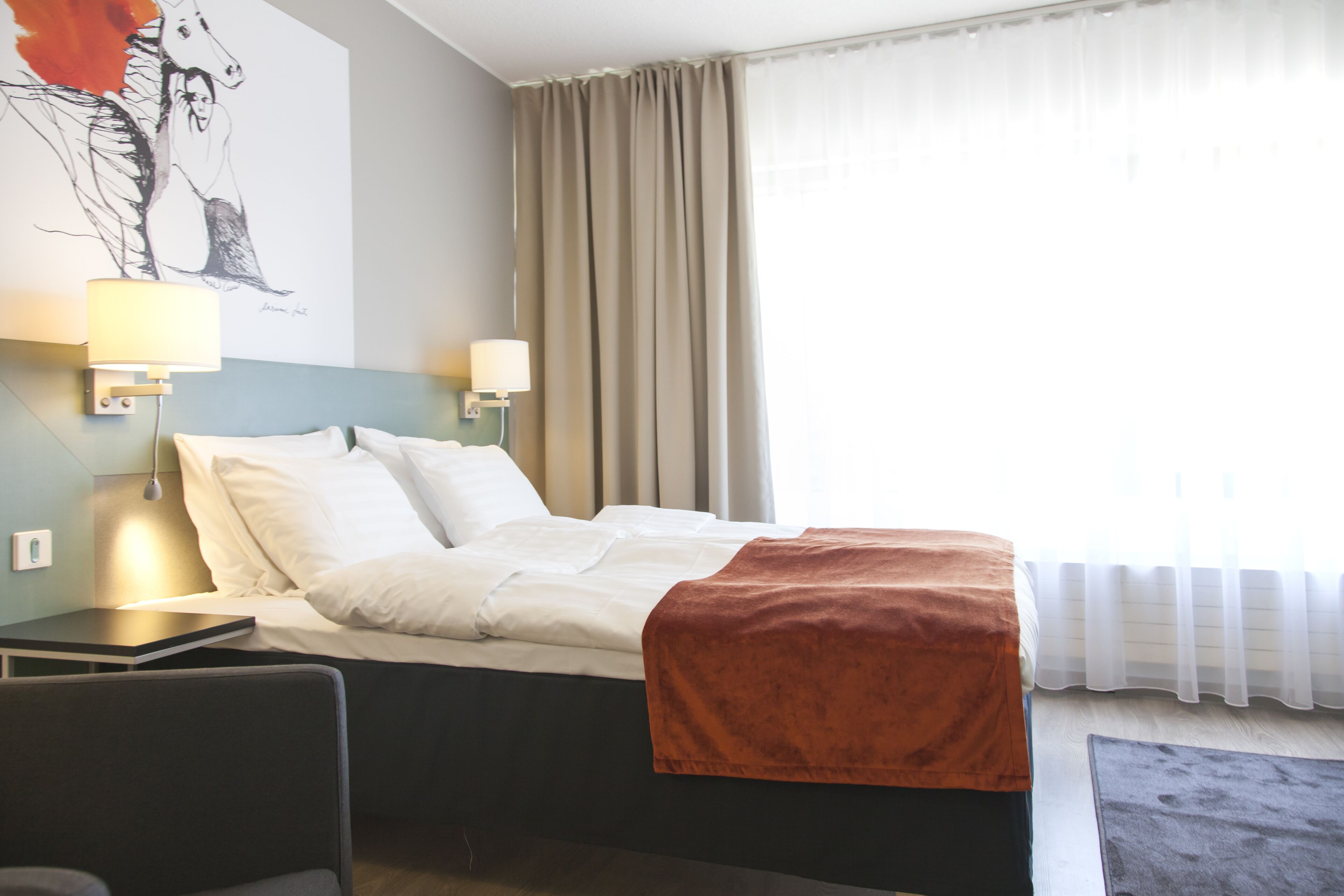 Hotellihuone, jossa on sänky, kaksi yöpöytävalaisinta, yöpöytä, työpöytä ja suuri ikkuna, jossa on verhot.