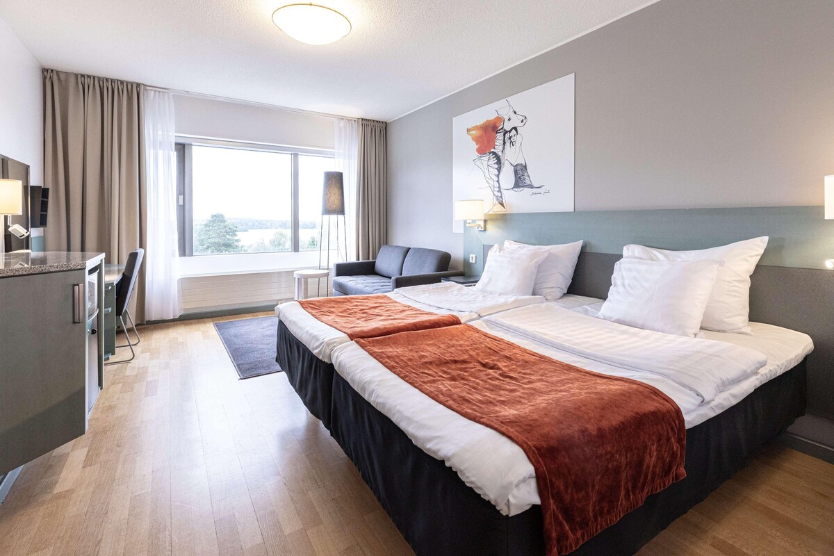 Una habitación de hotel con cama, escritorio, silla, ventana con cortinas y un cuadro en la pared.