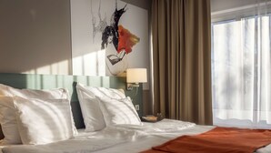 Habitación de hotel con cama, ropa de cama blanca, manta marrón, una obra de arte en la pared, una lámpara y una ventana con cortinas.
