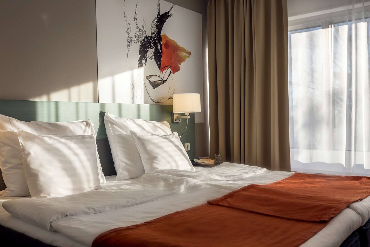 A hotel room with a bed, white bedding, a brown throw blanket, a wall art piece, a lamp, and a window with curtains.