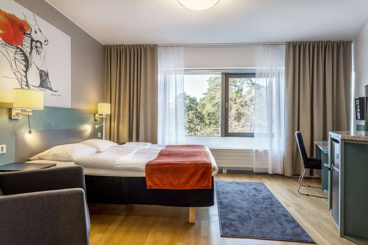 A modern hotel room with a bed, a desk, a chair, a window with curtains, and a painting on the wall.