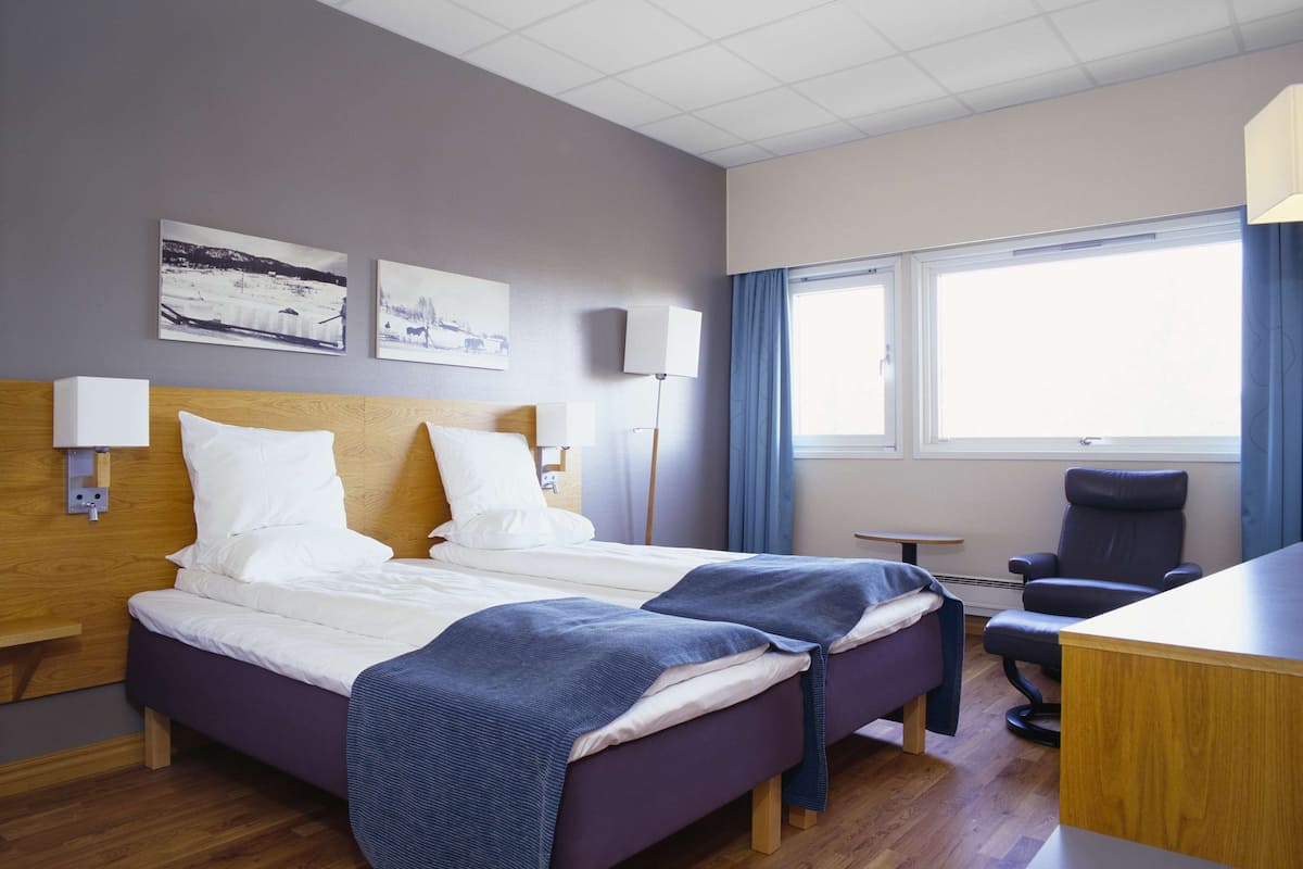 Una habitación de hotel con dos camas, un escritorio, una silla, una lámpara y una ventana con cortinas.