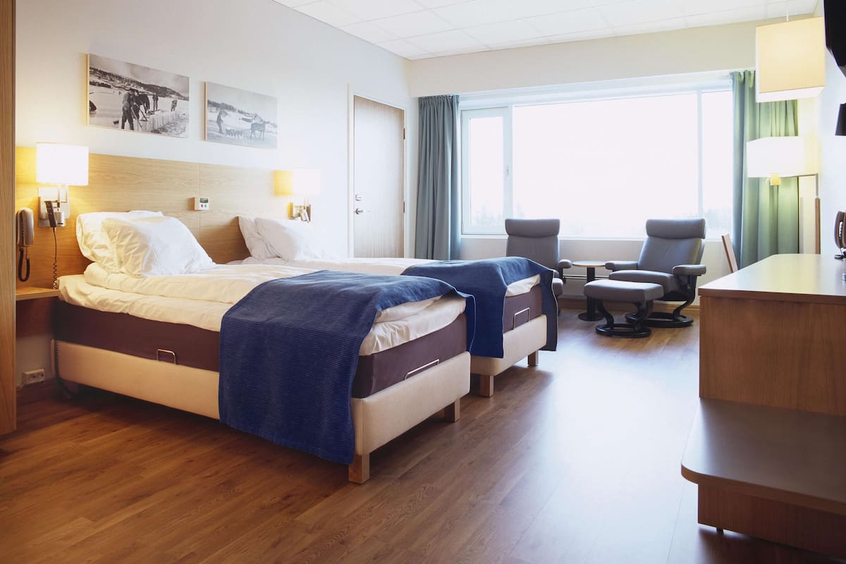 Una habitación de hotel con dos camas, un escritorio, una silla y un ventanal con cortinas.