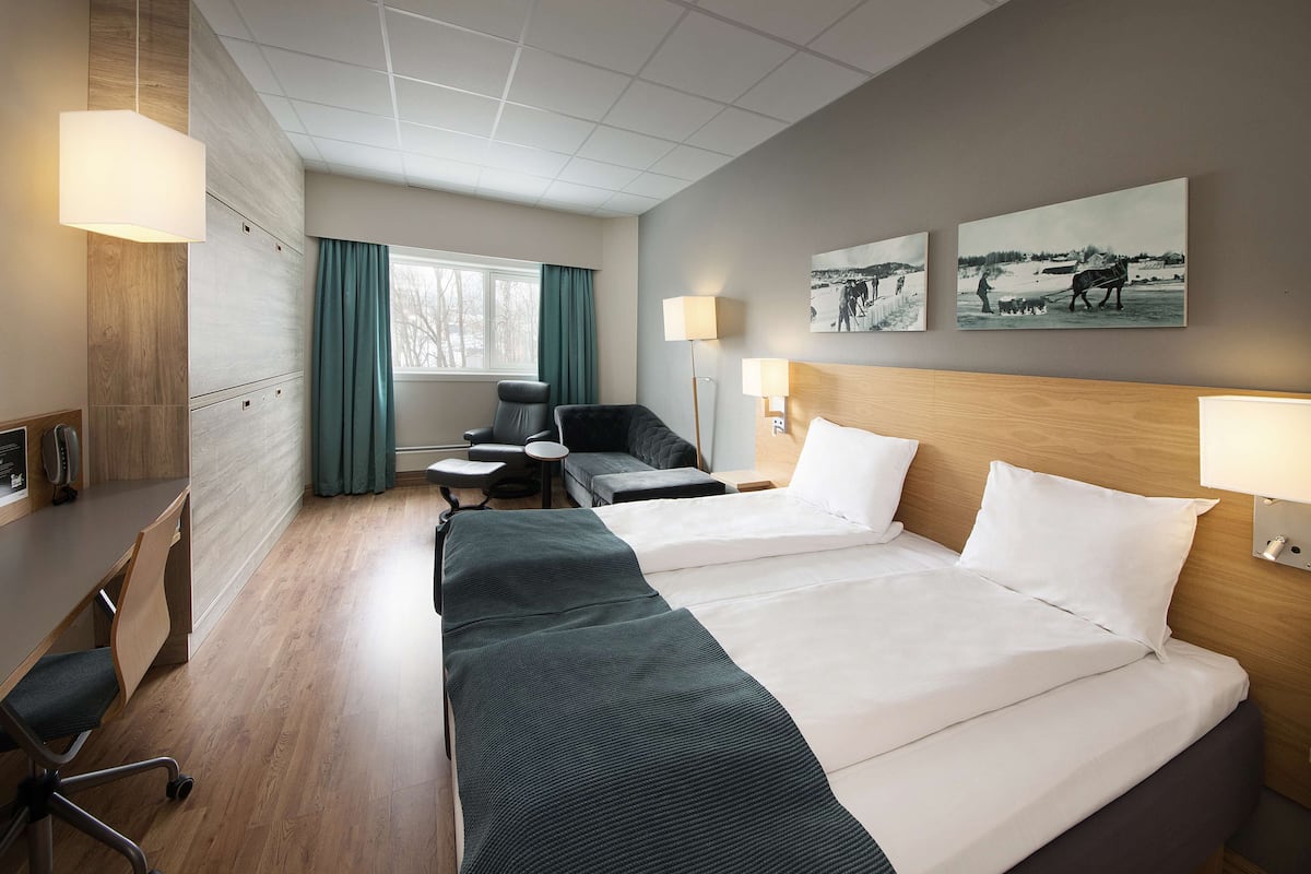Habitación de hotel con una cama grande, un escritorio, una silla, una lámpara y dos lámparas de pared. Hay dos cuadros en la pared.