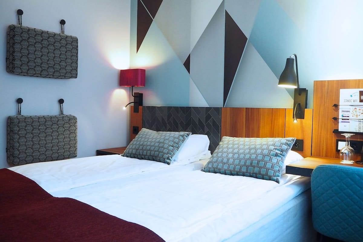 A hotel room with a bed, two nightstands, a desk, and a chair. There are wall-mounted lamps and a bulletin board with a sign.
