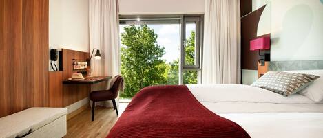 A hotel room with a bed, a desk, a chair, and a window with a view of trees.