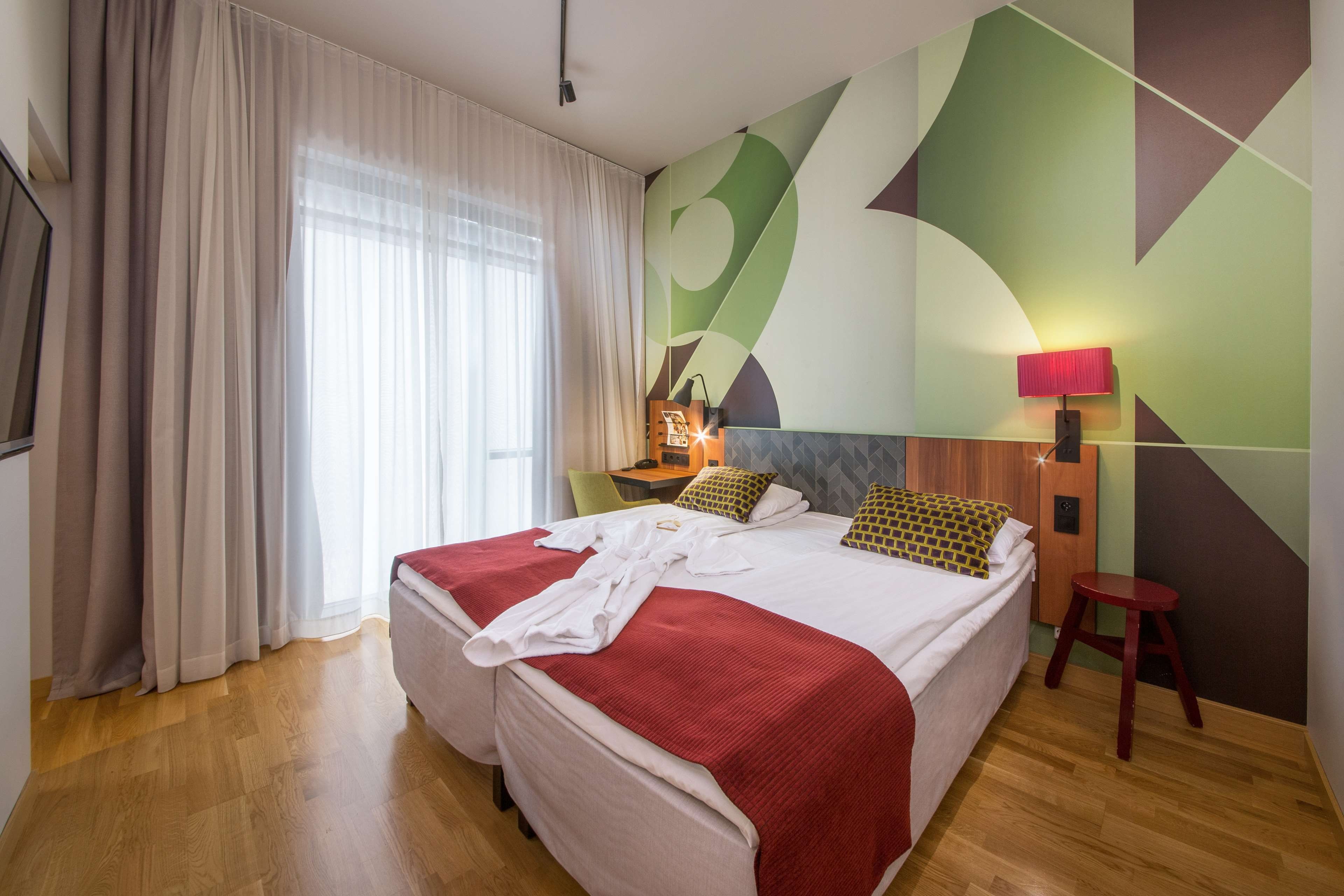 Habitación de hotel con dos camas, piso de madera, pared con patrones geométricos, lámpara roja y ventana con cortinas.