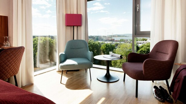 A modern hotel room with a large window, a round table, two chairs, a bed, and a red quilted chair. - Scandic Solli Oslo (Oslo)