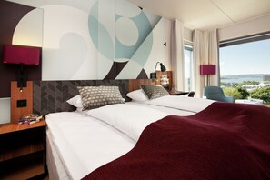 A hotel room with a large bed, two bedside tables, a nightstand, a desk, a chair, and a large window with a view. - Scandic Solli Oslo (Oslo)