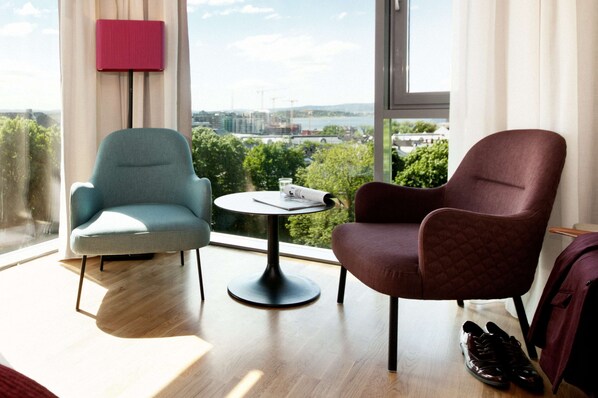 Ein modernes Zimmer mit zwei Sessel, einem kleinen Tisch und Blick auf eine Stadtlandschaft durch große Fenster.