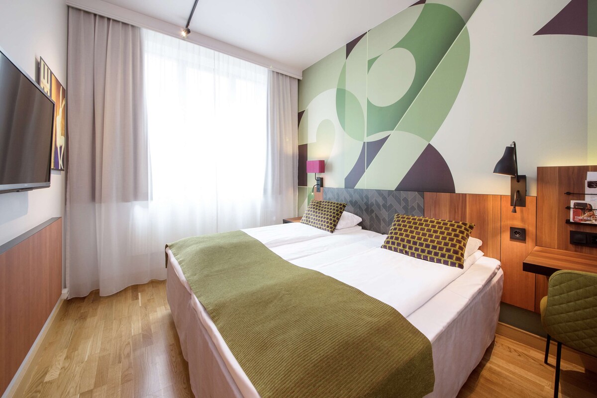 A hotel room with a large bed, a flat-screen TV, a desk with a lamp, and a green chair.