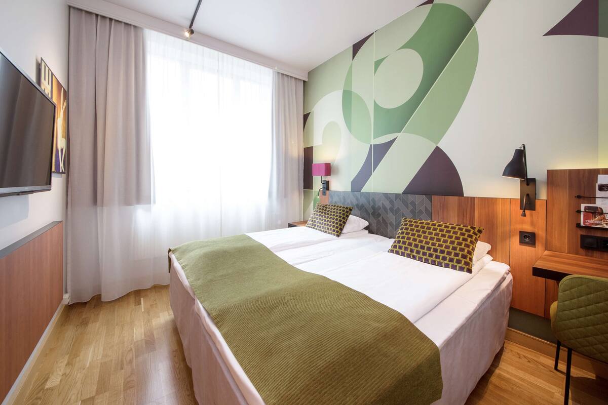 A hotel room with a large bed, a flat-screen TV, a desk with a lamp, and a green chair.