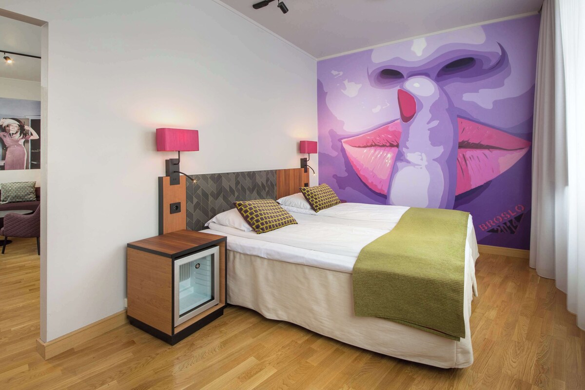 Una habitación de hotel moderna con una cama grande, dos lámparas de noche, una mesita de noche pequeña y un mural en la pared.