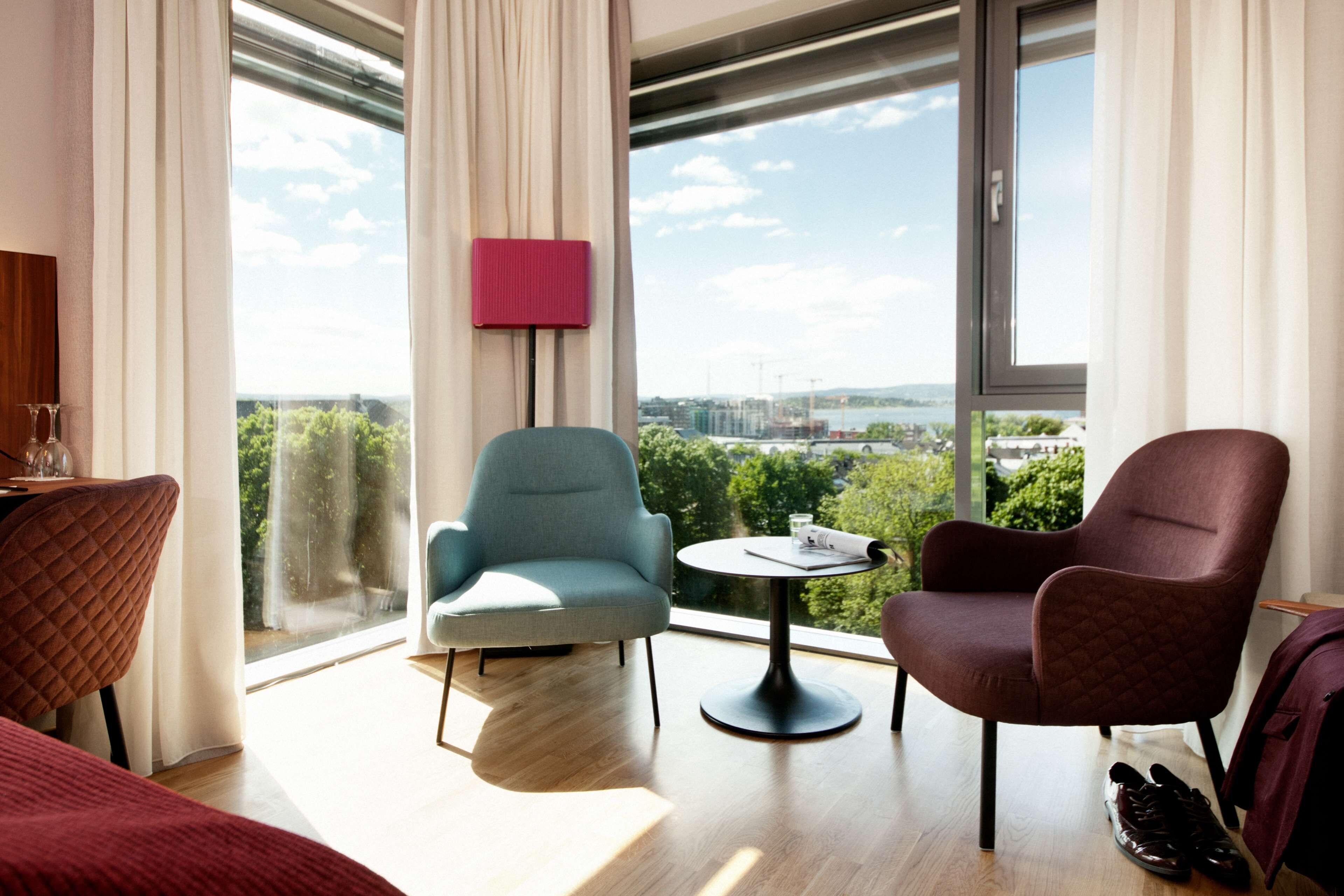 Une chambre d’hôtel moderne avec une grande fenêtre, une table ronde, deux chaises, un lit et un fauteuil rouge matelassé.
