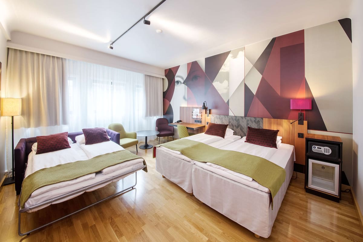 A hotel room with two beds, a desk, a chair, a lamp, and a microwave.