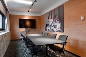 Una sala conferenze con un lungo tavolo, sedie, un orologio, un televisore e un quadro di grandi dimensioni appeso al muro.