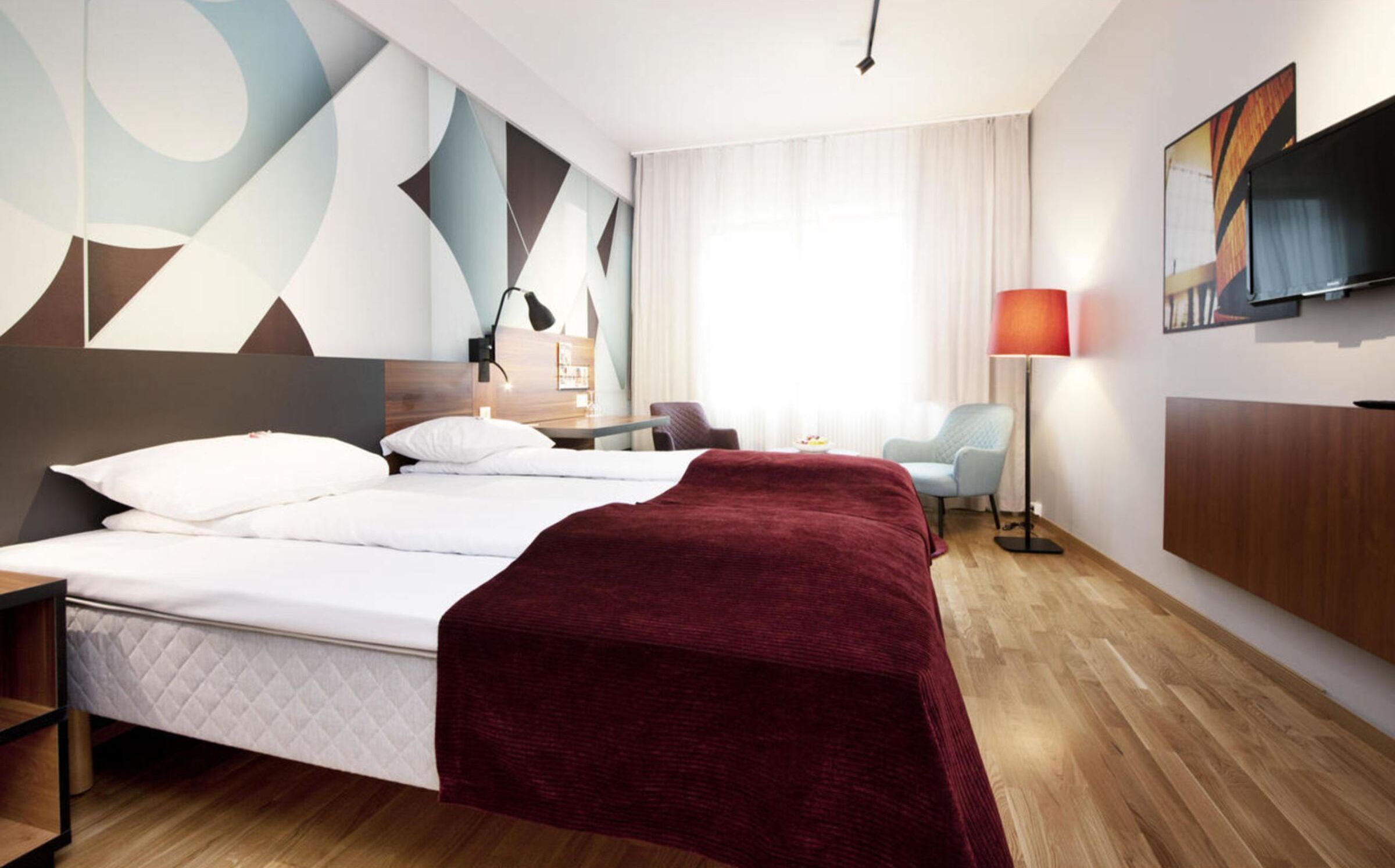 A modern hotel room with a large bed, a desk, a chair, a lamp, and a painting on the wall.