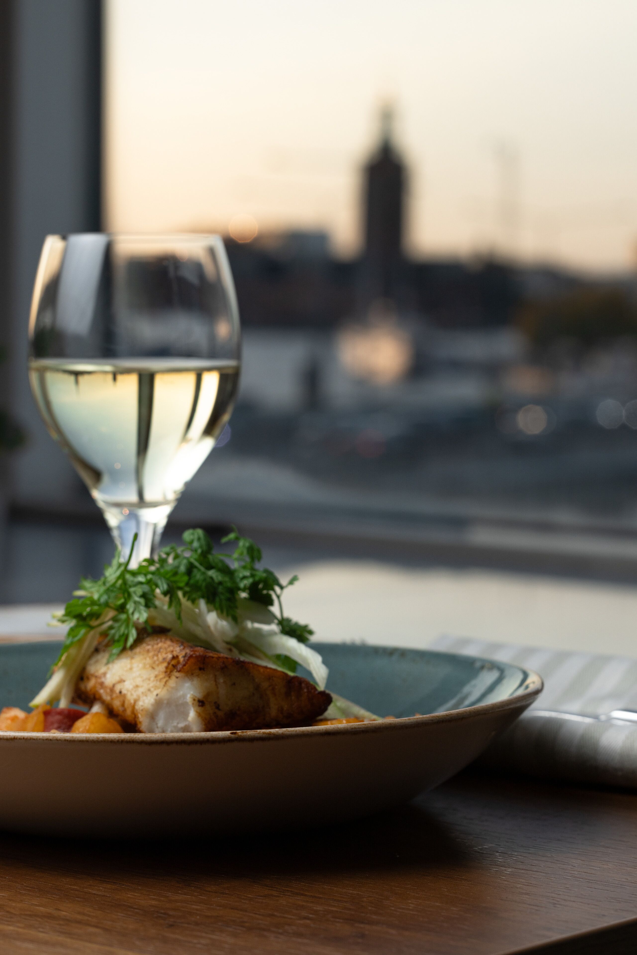 A plate of food with a glass of white wine on a table by a window with a cityscape view.