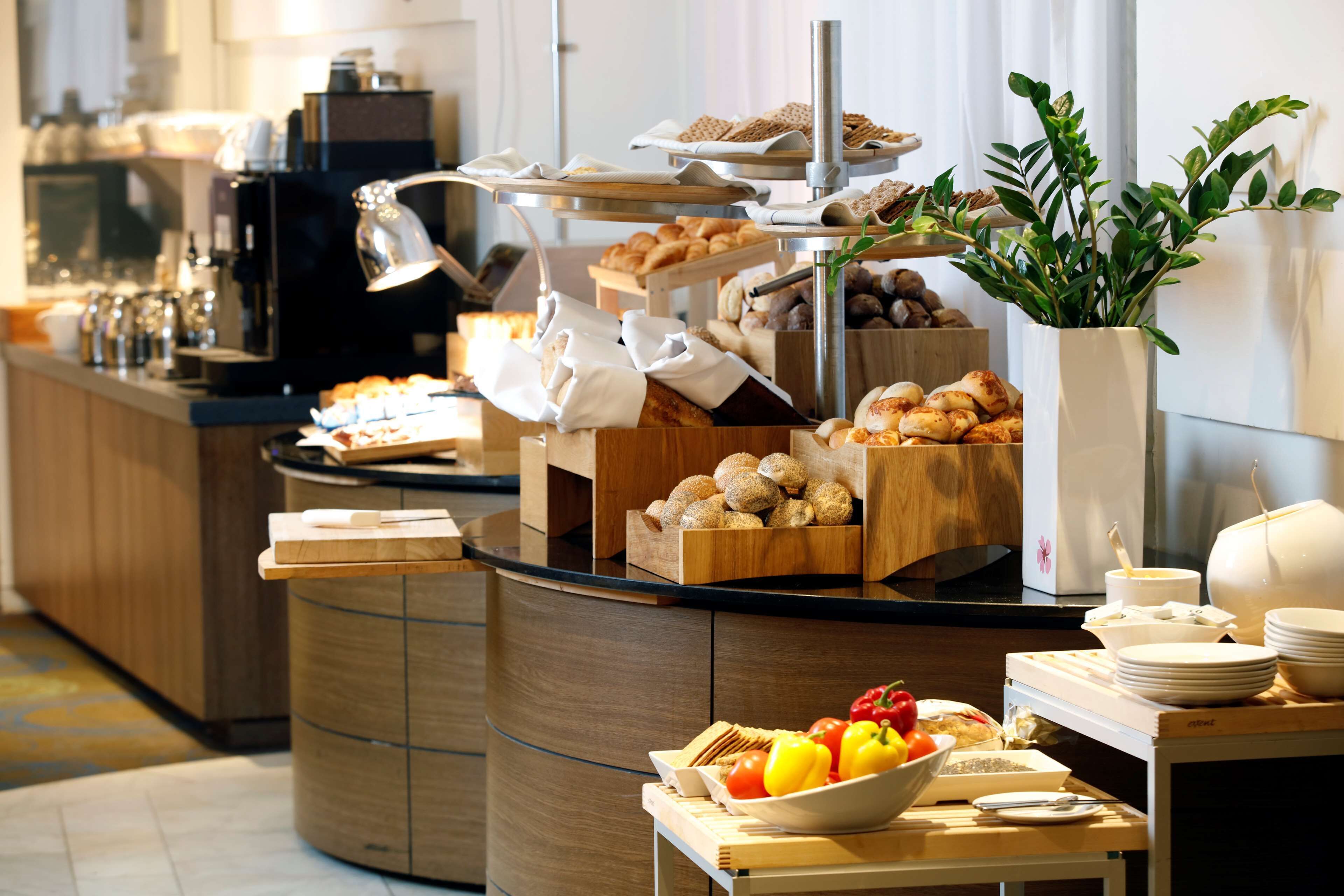 A buffet setup with various food items, including fruits, pastries, and bread, arranged on a counter with a coffee machine and a plant in the background.