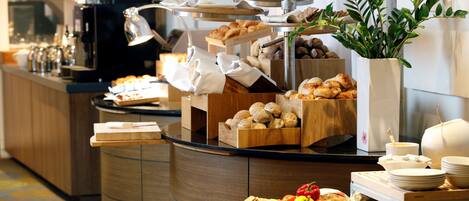 A buffet setup with various food items, including fruits, pastries, and bread, arranged on a counter with a coffee machine and a plant in the background.