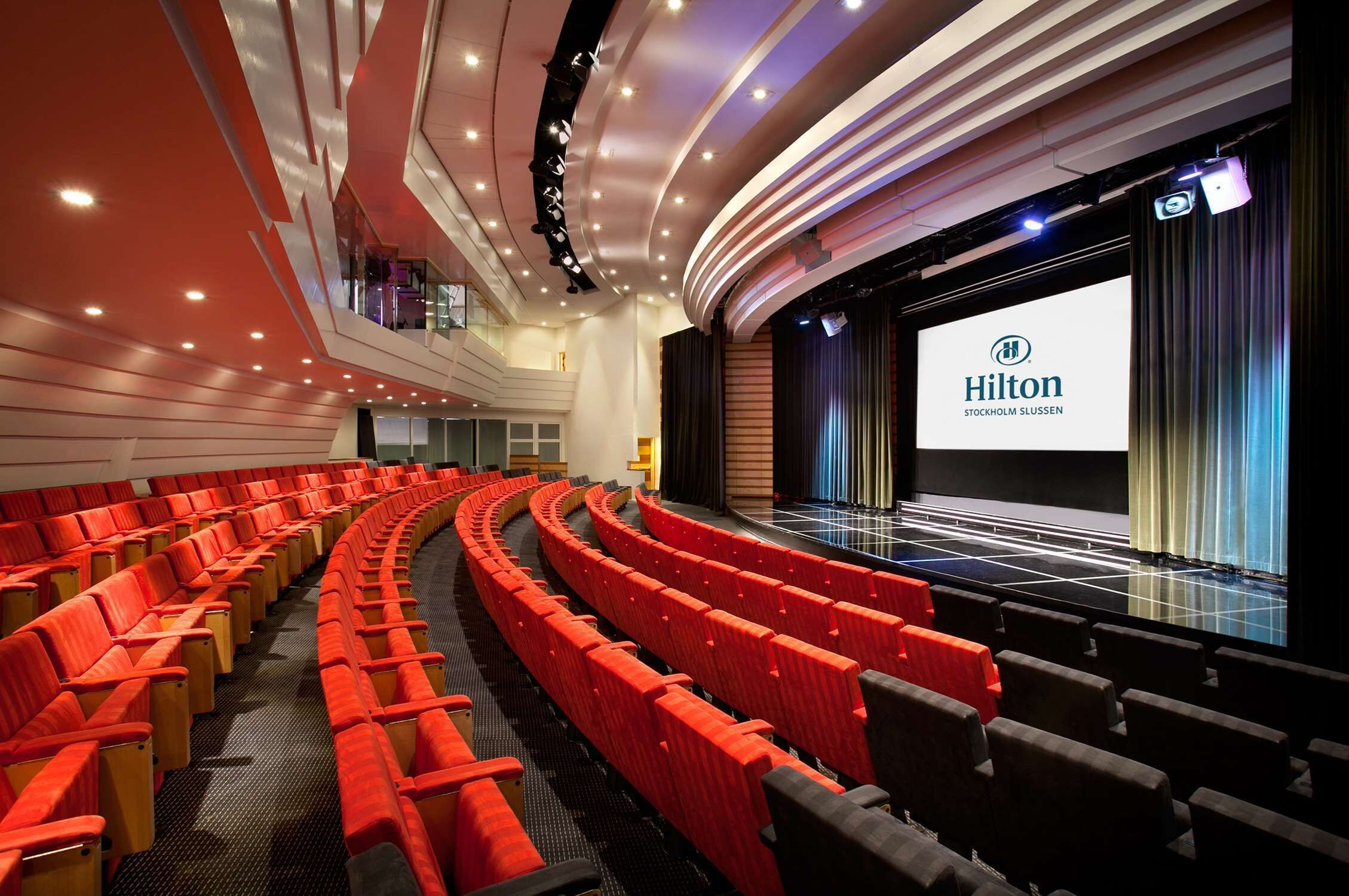 Une salle de théâtre avec des sièges rouges, une scène et un écran affichant le texte « Hilton Stockholm Slussen ».