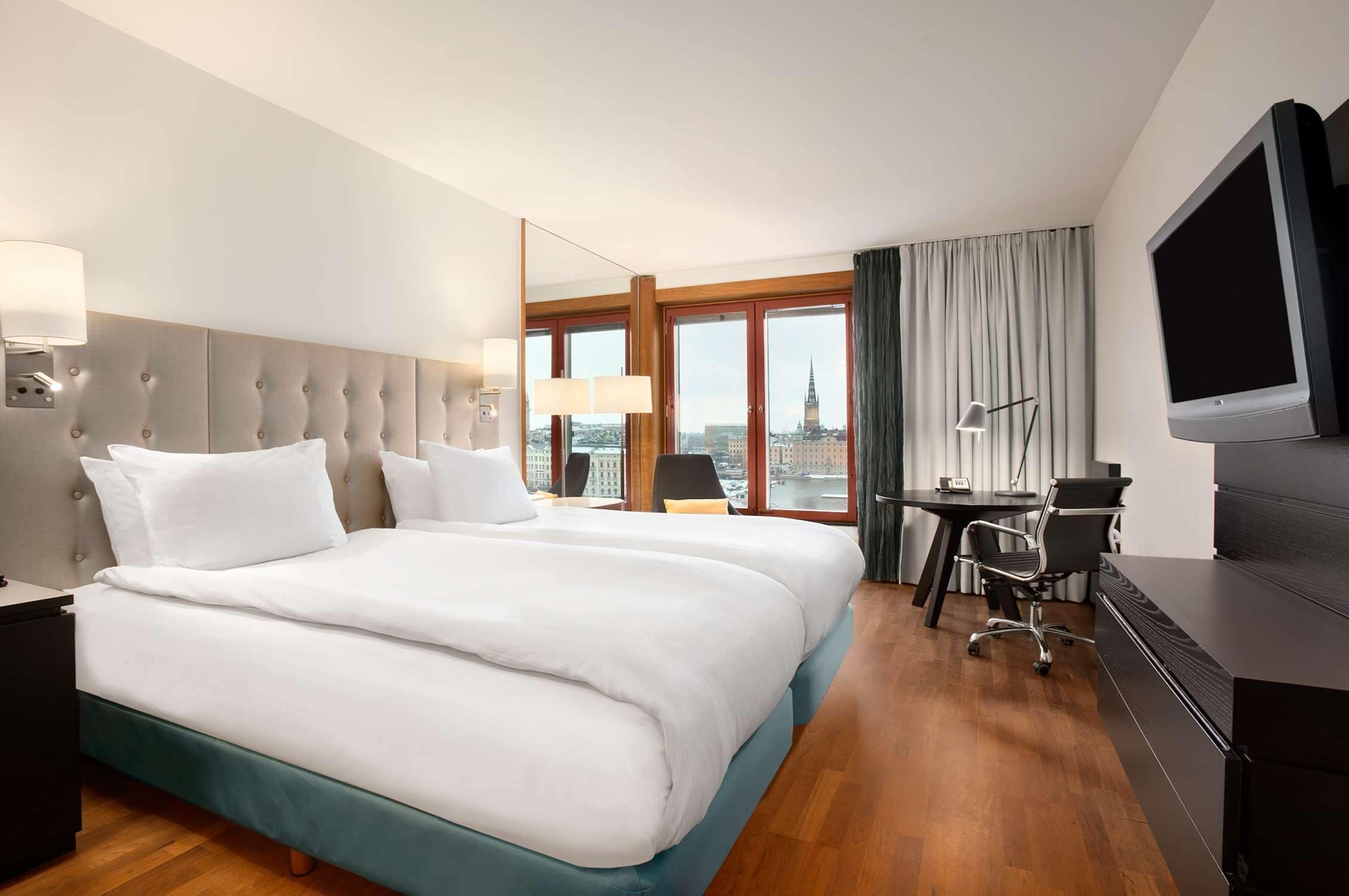 A hotel room with a large bed, a desk, a chair, a TV, and a city view.