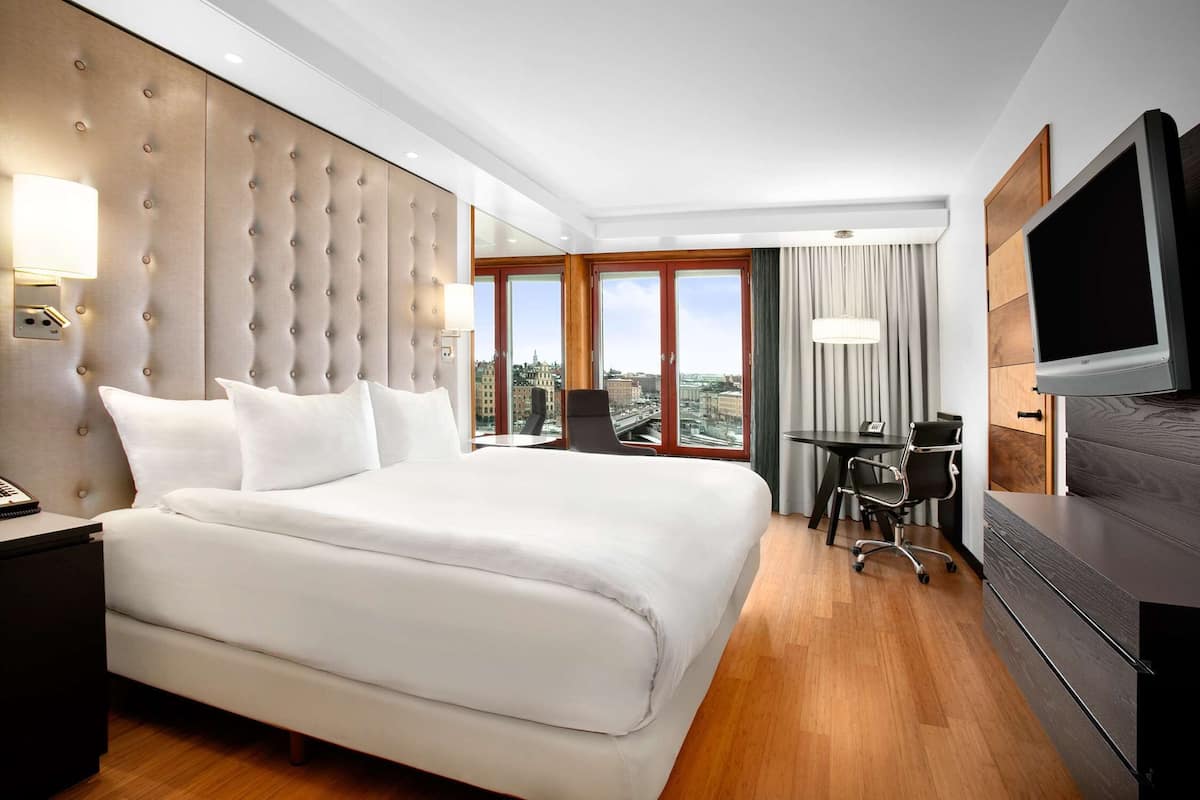 A hotel room with a large bed, a desk, a chair, a television, and a view of the city through large windows.