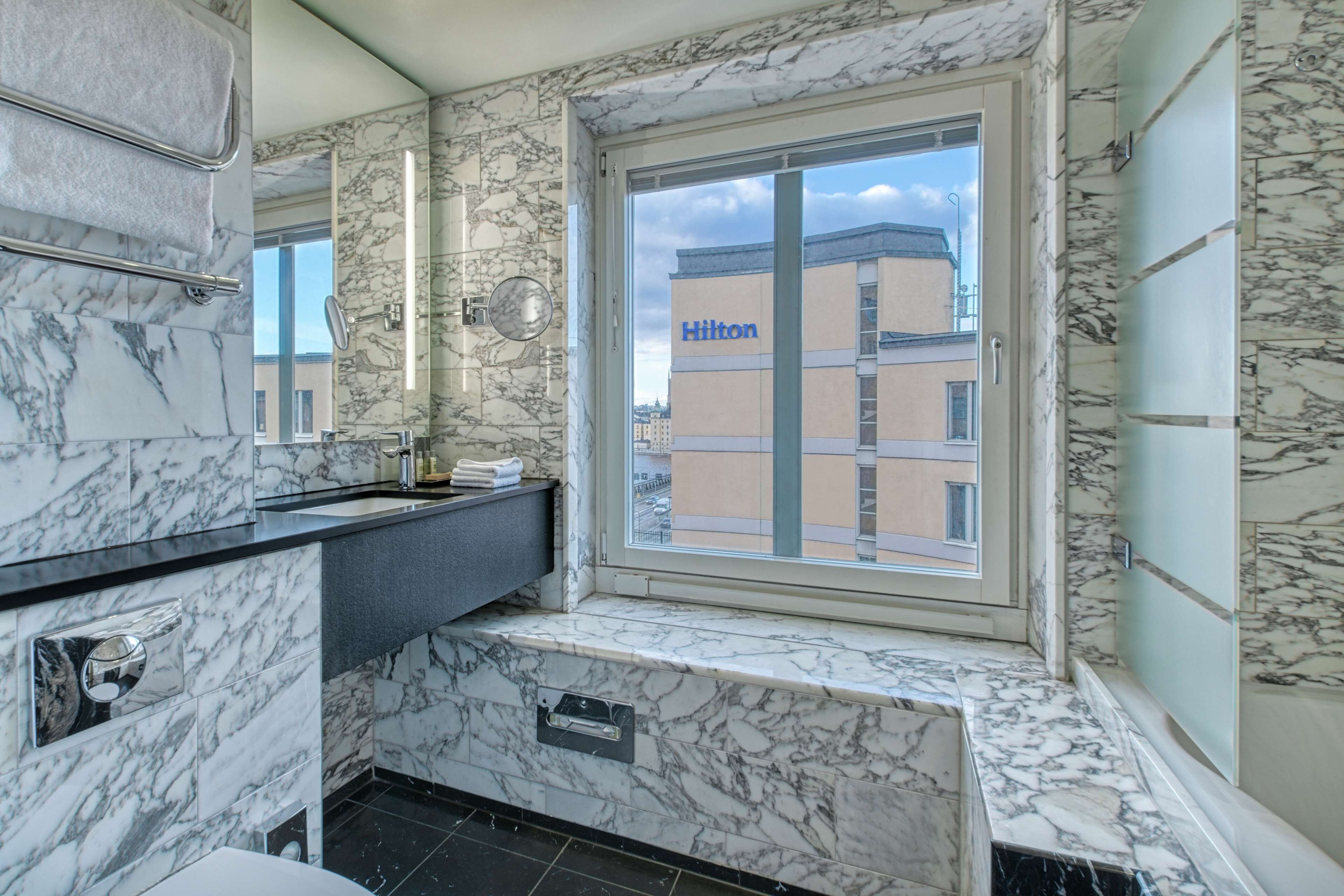 Un baño moderno con paredes de mármol, un ventanal y vista a un edificio Hilton.