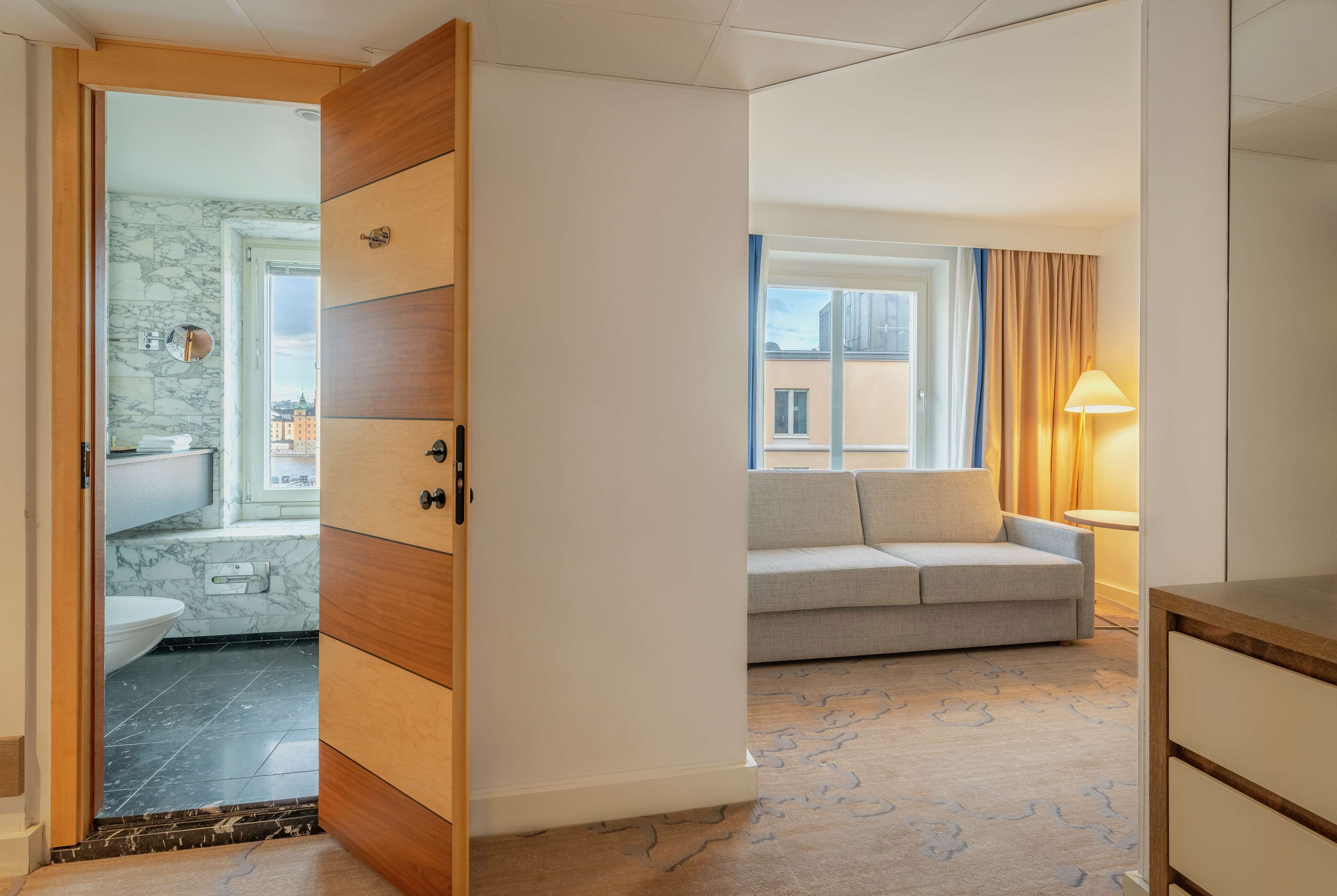 A modern hotel room with a wooden wardrobe, a grey sofa, a window with curtains, and a view of buildings outside.