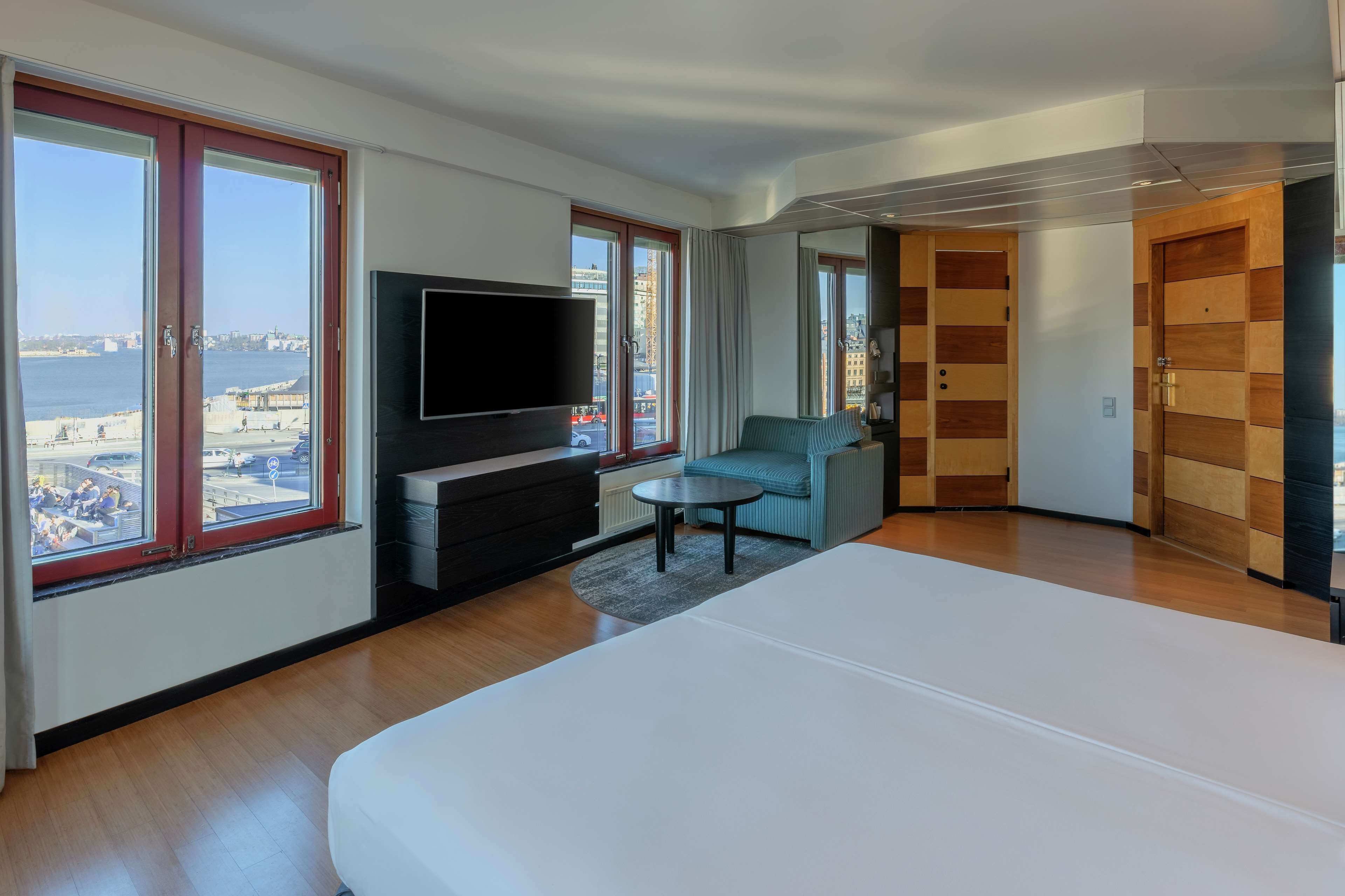 A modern hotel room with a large bed, a flat-screen TV, a seating area with a sofa and armchair, and a view of the city and sea through large windows.
