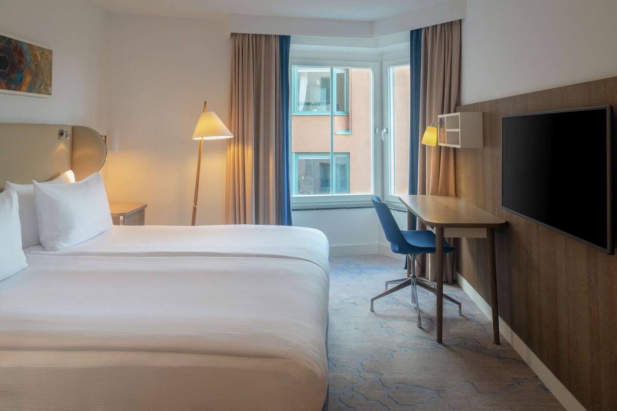 A hotel room with a large bed, a desk, a chair, a flat-screen TV, a lamp, and a window with curtains.