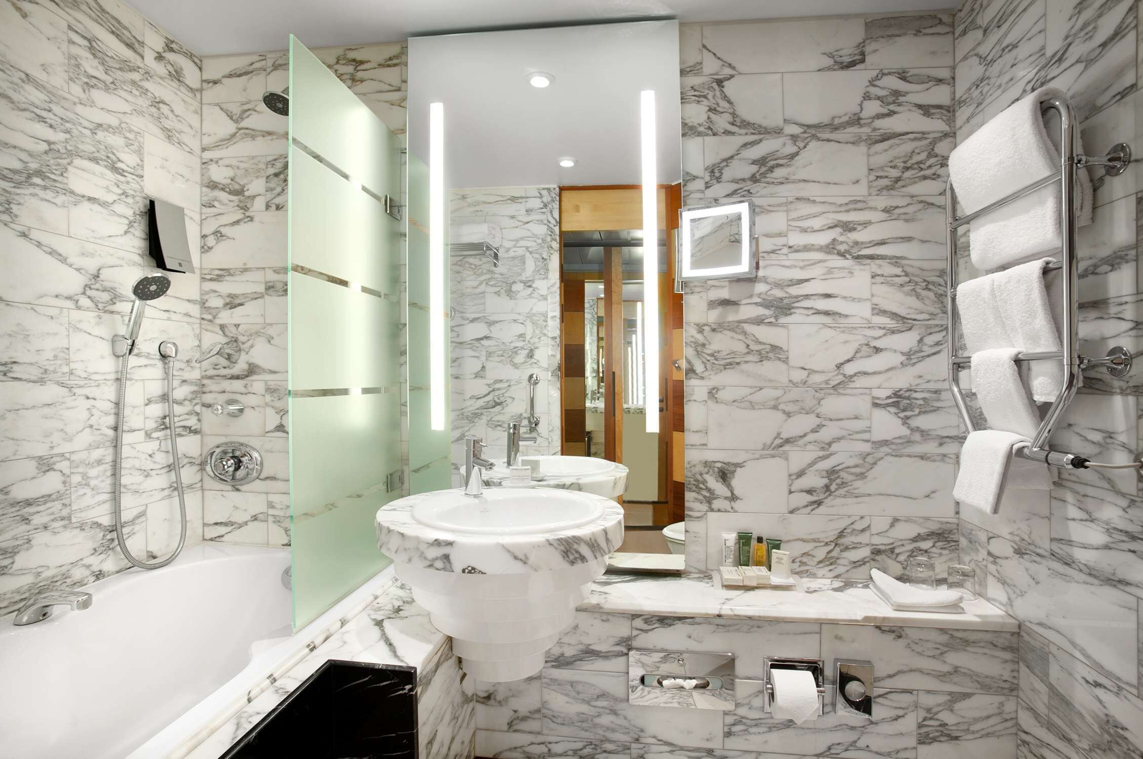A modern bathroom with marble walls, a bathtub, two sinks, and a glass shower enclosure.